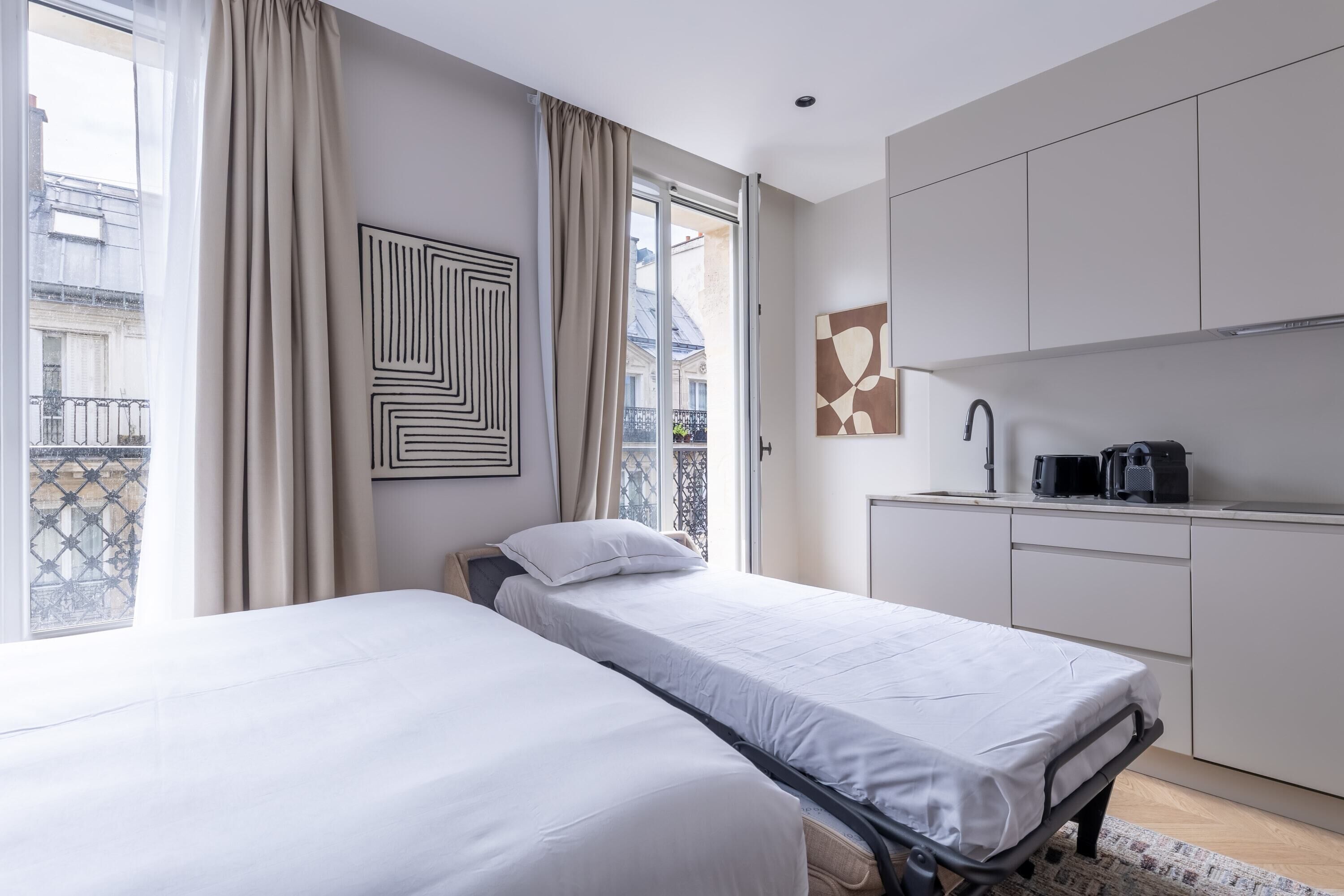 A modern hotel room with two beds, a kitchenette, and a balcony with a view of the city.
