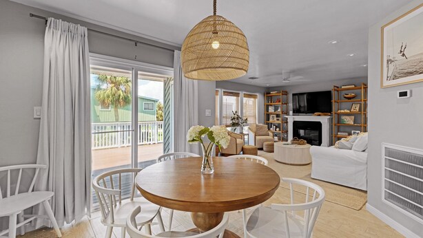 A modern living room with a round wooden table, white chairs, a hanging wicker light fixture, a white sofa, a fireplace, and a balcony with a view of palm trees. - Pirate's Beach Retreat! A short walk to the beach. (Galveston)