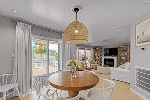 A modern living room with a round wooden table, white chairs, a hanging wicker light fixture, a white sofa, a fireplace, and a balcony with a view of palm trees. - Pirate's Beach Retreat! A short walk to the beach. (Galveston)