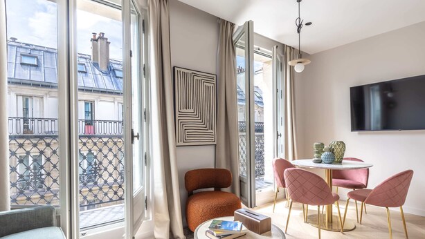 A modern living room with a large window, a round table with chairs, a sofa, and a mounted TV.