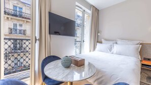 Une chambre d’hôtel avec un lit, un téléviseur fixé au mur, une table ronde avec un vase, et une vue sur la ville par la fenêtre.