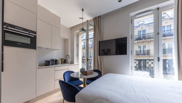 Une chambre d’hôtel moderne comprenant un lit, une table à manger avec des chaises, un coin cuisine et un balcon offrant une vue sur la ville.