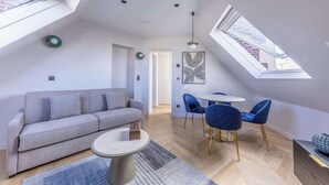 Un appartement moderne de style « loft », doté d’un plafond haut, de fenêtres de toit, d’un canapé gris, d’une table basse ronde et d’un coin repas avec des chaises bleues.