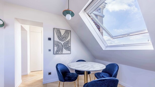 Un espace repas moderne avec une table ronde et des chaises en velours bleu, une verrière et une œuvre d’art abstraite encadrée accrochée au mur.