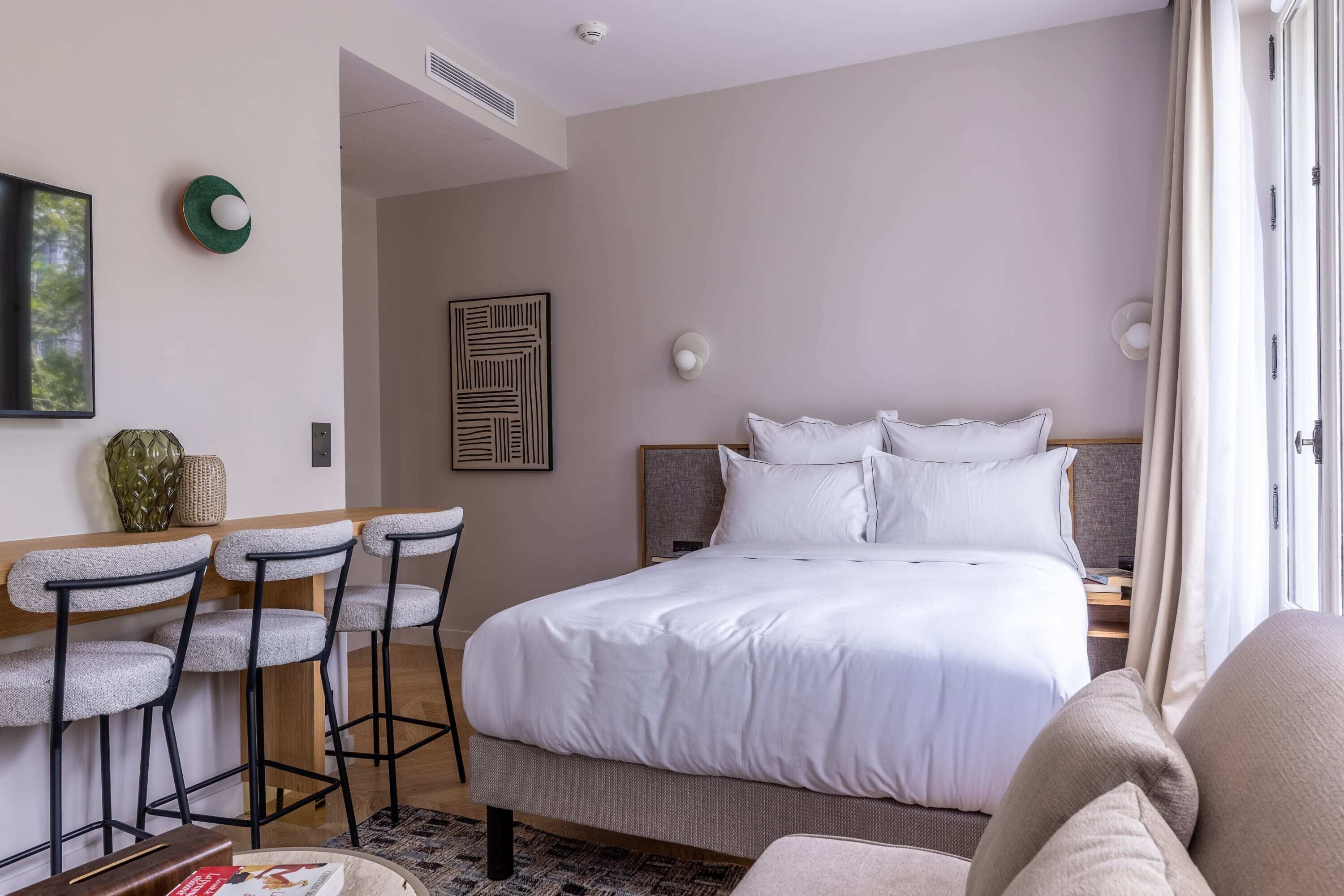 Hotelkamer met een groot bed, een bureau met twee barkrukken, een kunstwerk aan de muur en een raam met gordijnen.