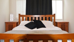 A bed with a wooden headboard, two black pillows, and white bedding. There are two wooden nightstands with a decorative item on one. A window with a black curtain is visible in the background.