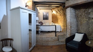 Ein Zimmer mit Steinwand, einem Bett mit weißer Bettdecke, einem schwarzen Stuhl, einem kleinen runden Tisch mit zwei Tassen und einem Schrank mit Spiegel.