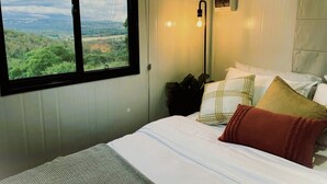 A bedroom with a large window offering a view of a green landscape. There is a bed with white bedding and multiple pillows. A wall-mounted light fixture is visible. - Tiny Home in Hampton (Hampton)