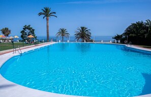 A clear blue swimming pool with a view of the ocean, surrounded by palm trees and umbrellas.