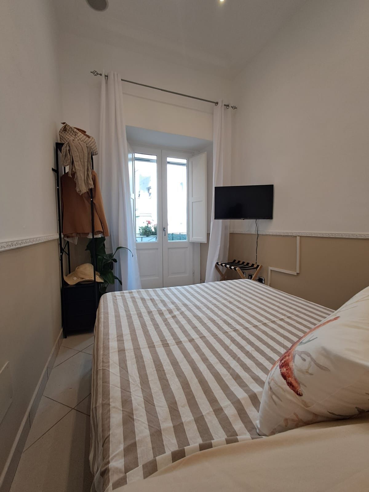 A bedroom with a bed, a TV mounted on the wall, a window with curtains, and a coat rack with clothes.