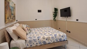 Egy hálószoba, amelyben van egy ágy, egy festmény, egy légkondicionáló, egy televízió és egy növény.