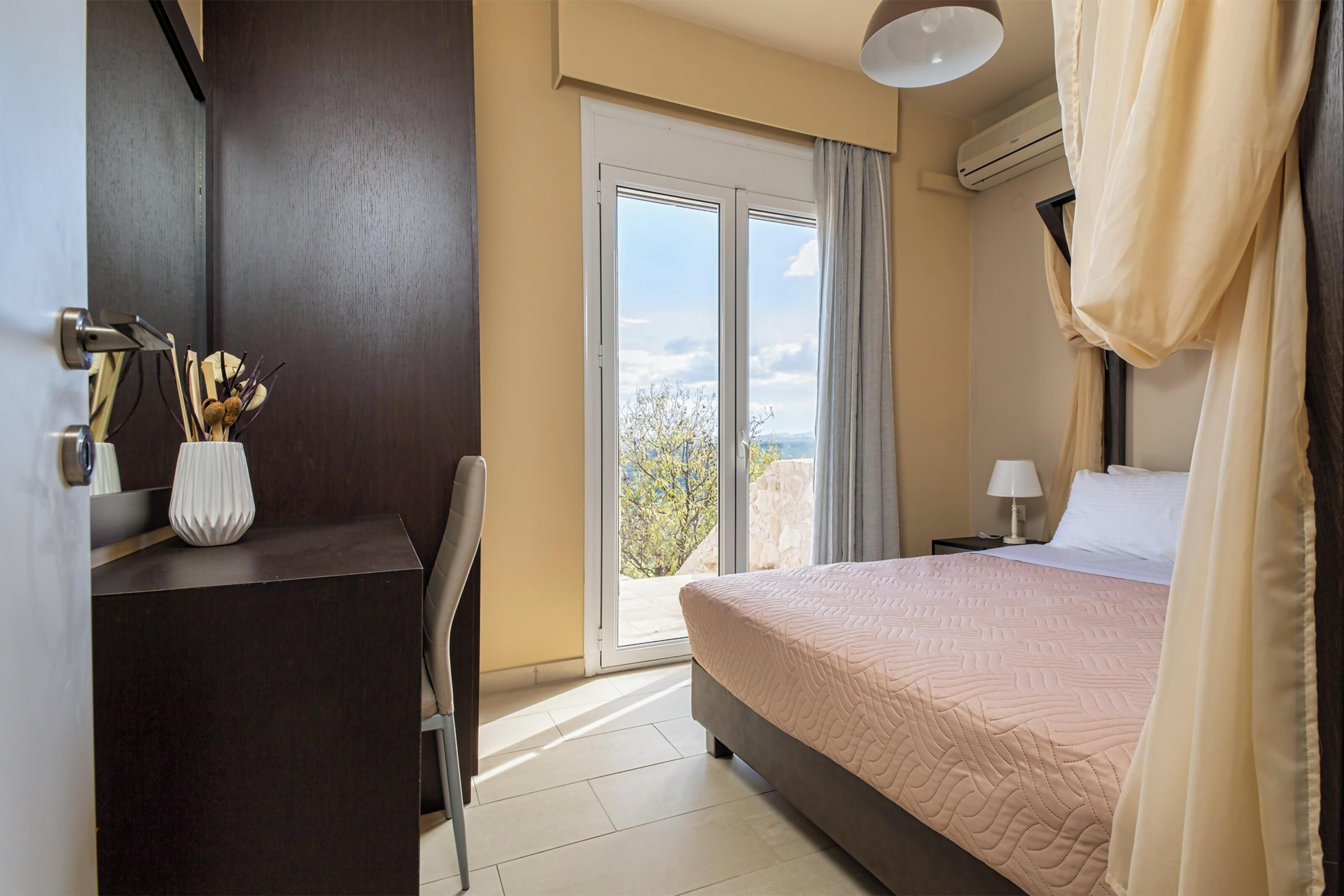 Una habitación de hotel con cama, escritorio, silla, mesita de noche con lámpara y vistas al exterior a través de una puerta corredera.