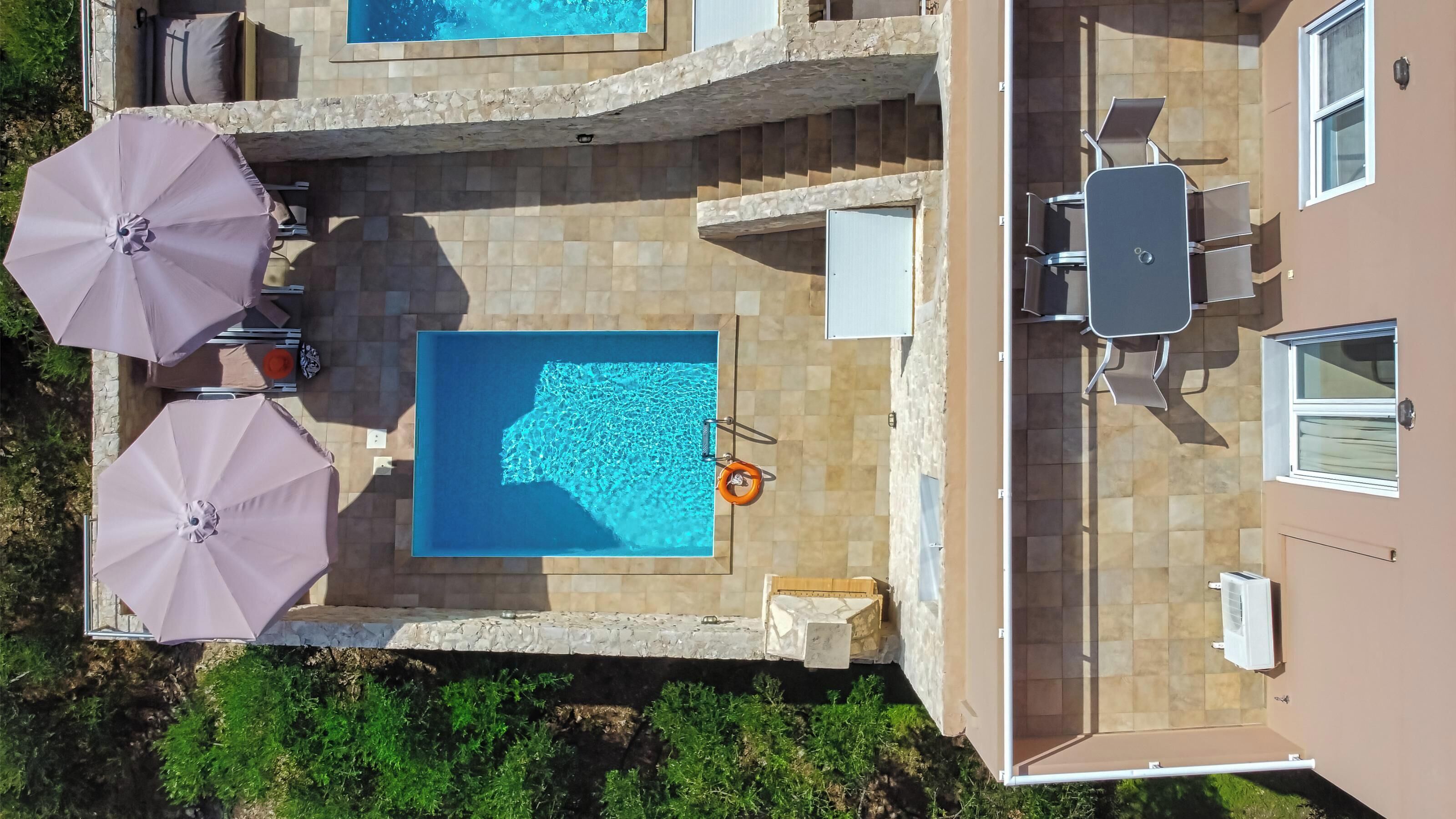 An aerial view of a rooftop pool area with two pools, surrounded by stone walls, and equipped with umbrellas and lounge chairs.