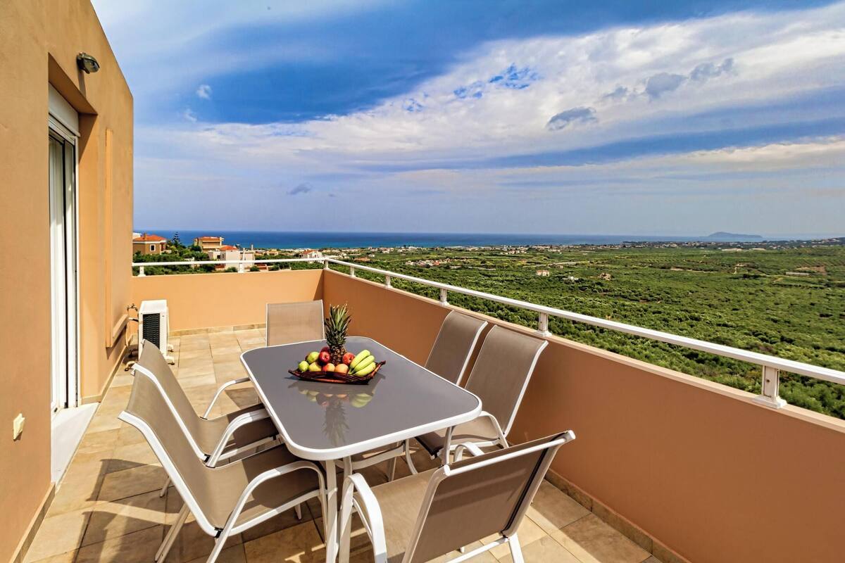 Un balcon avec une table dressée pour deux personnes, des chaises et un panier de fruits. Le balcon offre une vue sur un paysage verdoyant et la mer.