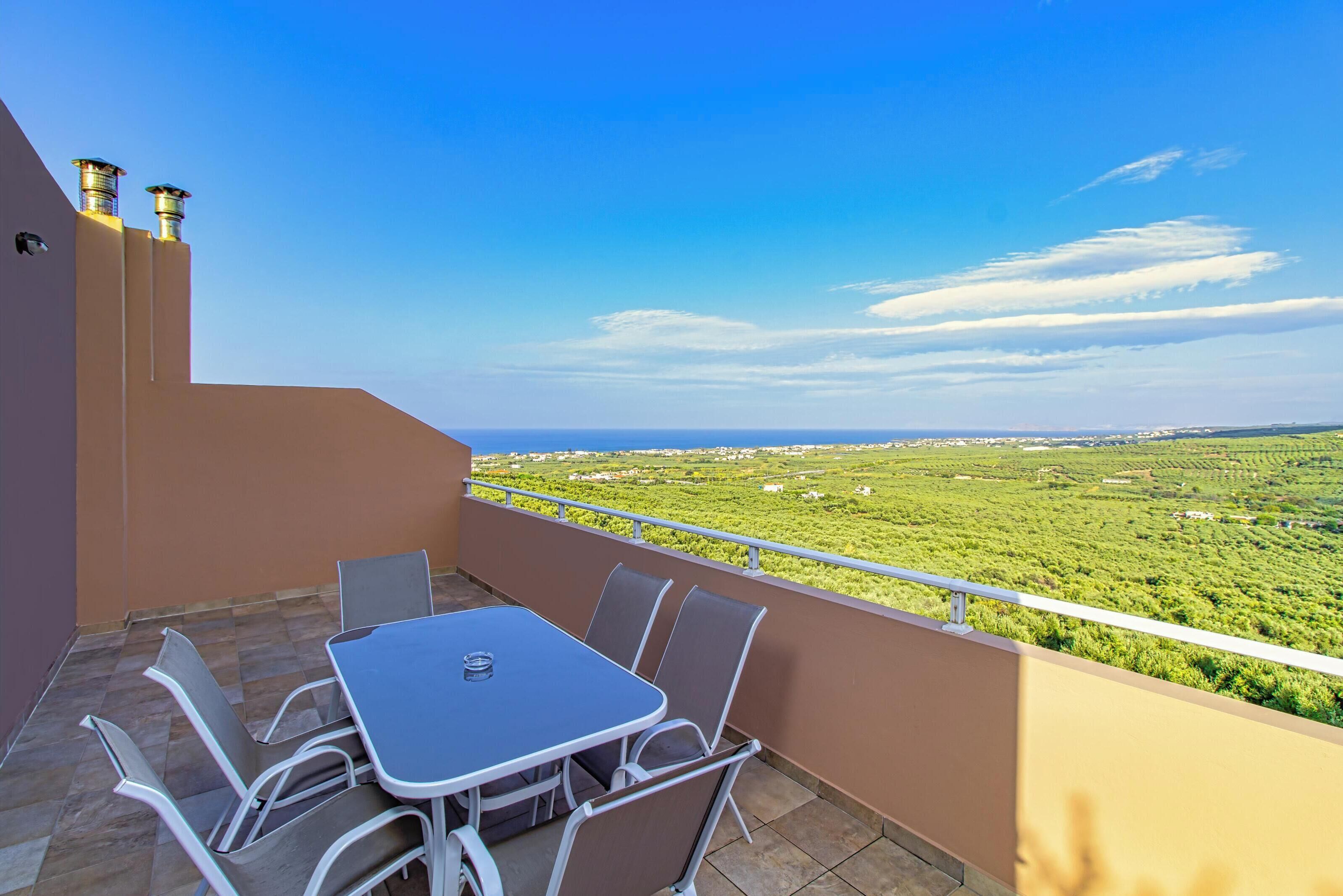 A balcony with a table and chairs, offering a view of a green landscape and distant buildings.