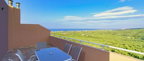 A balcony with a table and chairs, offering a view of a green landscape and distant buildings.