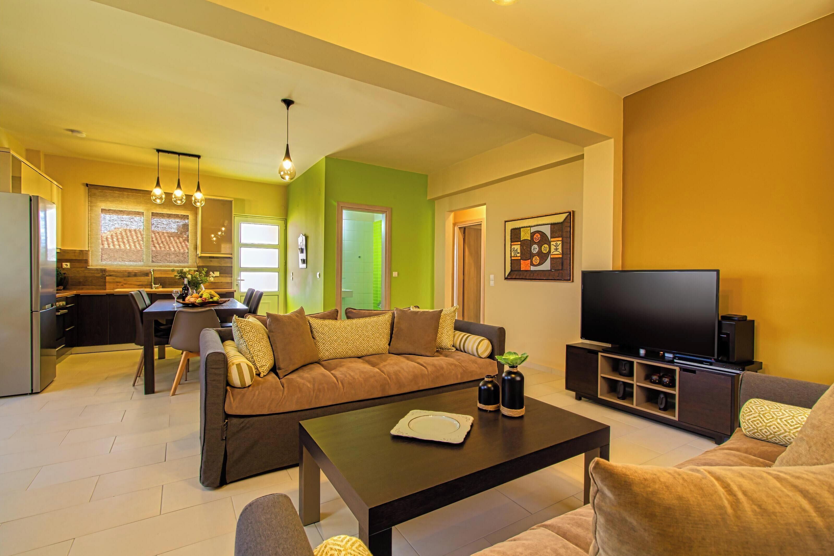 Una sala de estar moderna con sofá, mesa de centro y televisor. Se observa un comedor con mesa y sillas, y una cocina al fondo.
