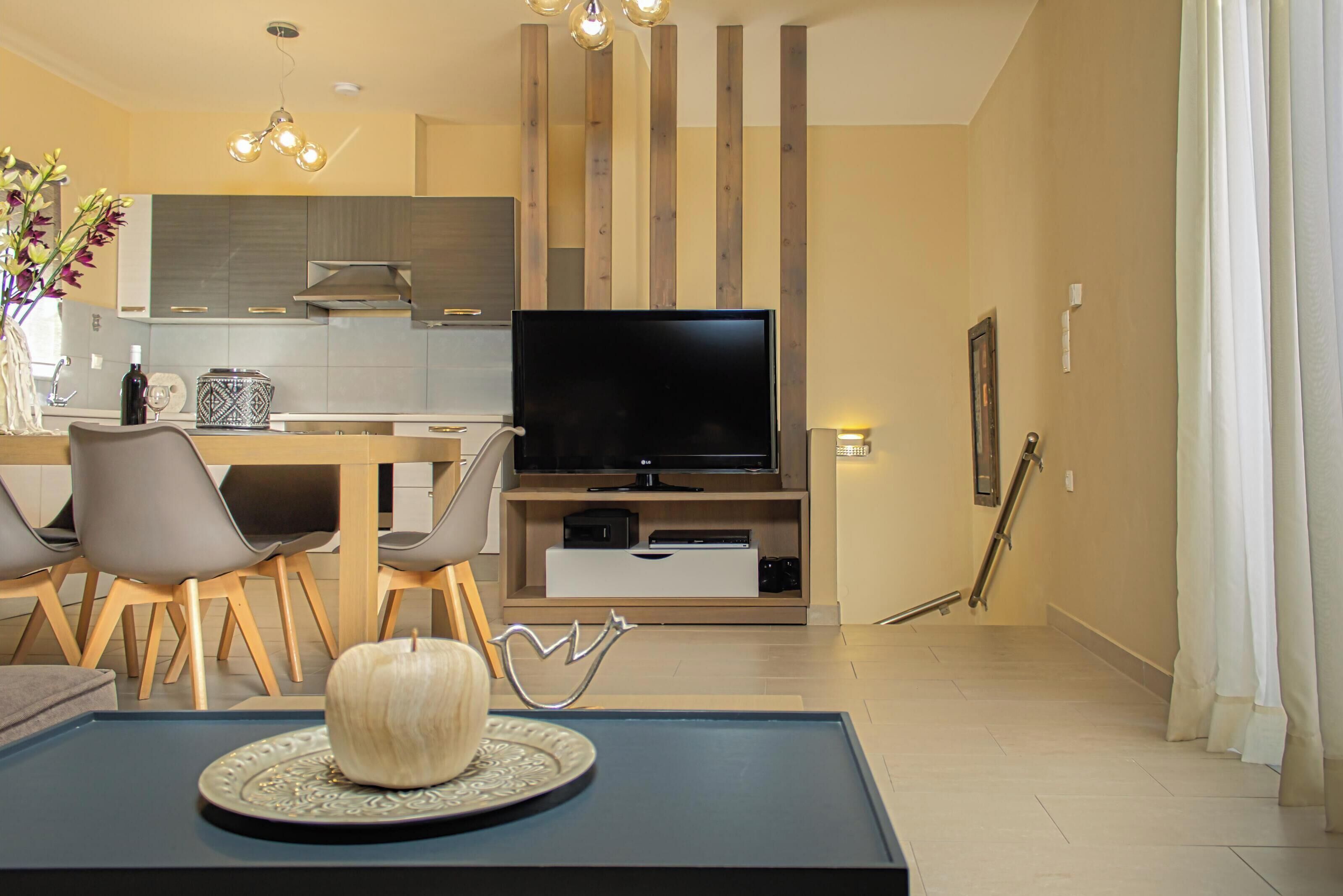 A modern living room with a flat-screen TV, a dining area with chairs, a coffee table with a decorative item, and a kitchen in the background.