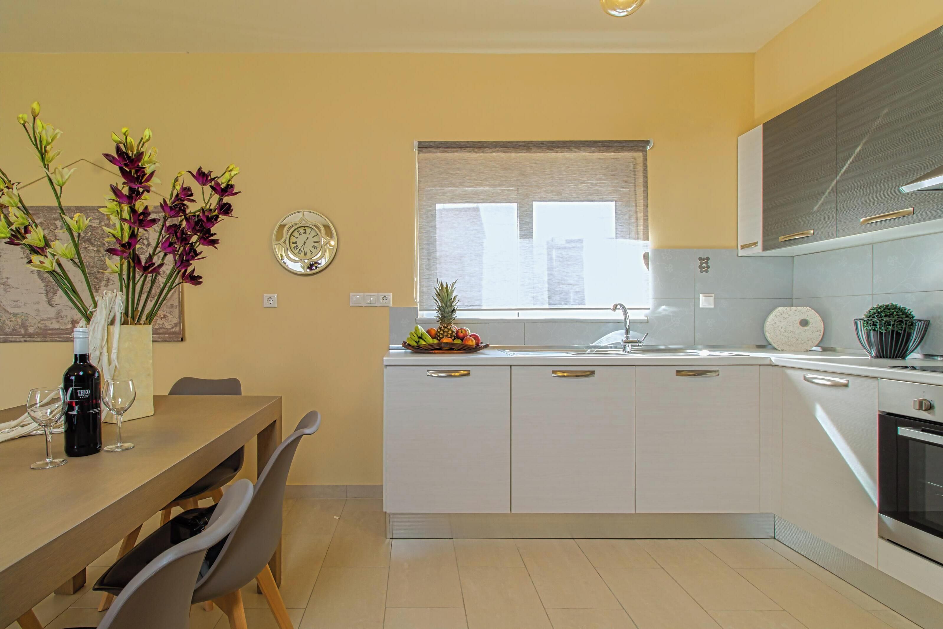 A modern kitchen with white cabinets, a dining table with chairs, a window with blinds, and a fruit bowl on the counter.