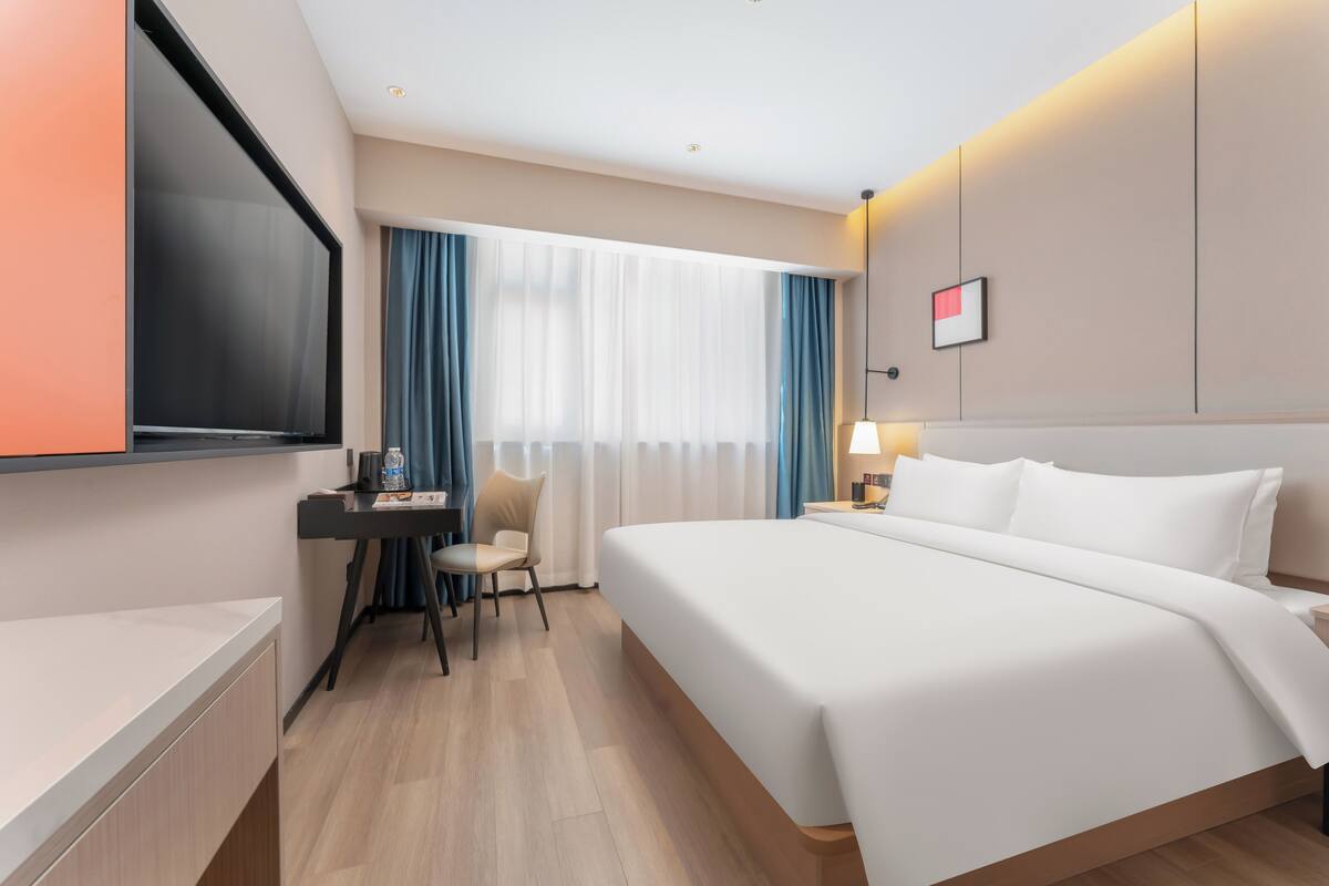 Habitación de hotel con una cama grande, un escritorio con silla, un televisor de pantalla plana y un ventanal con cortinas.