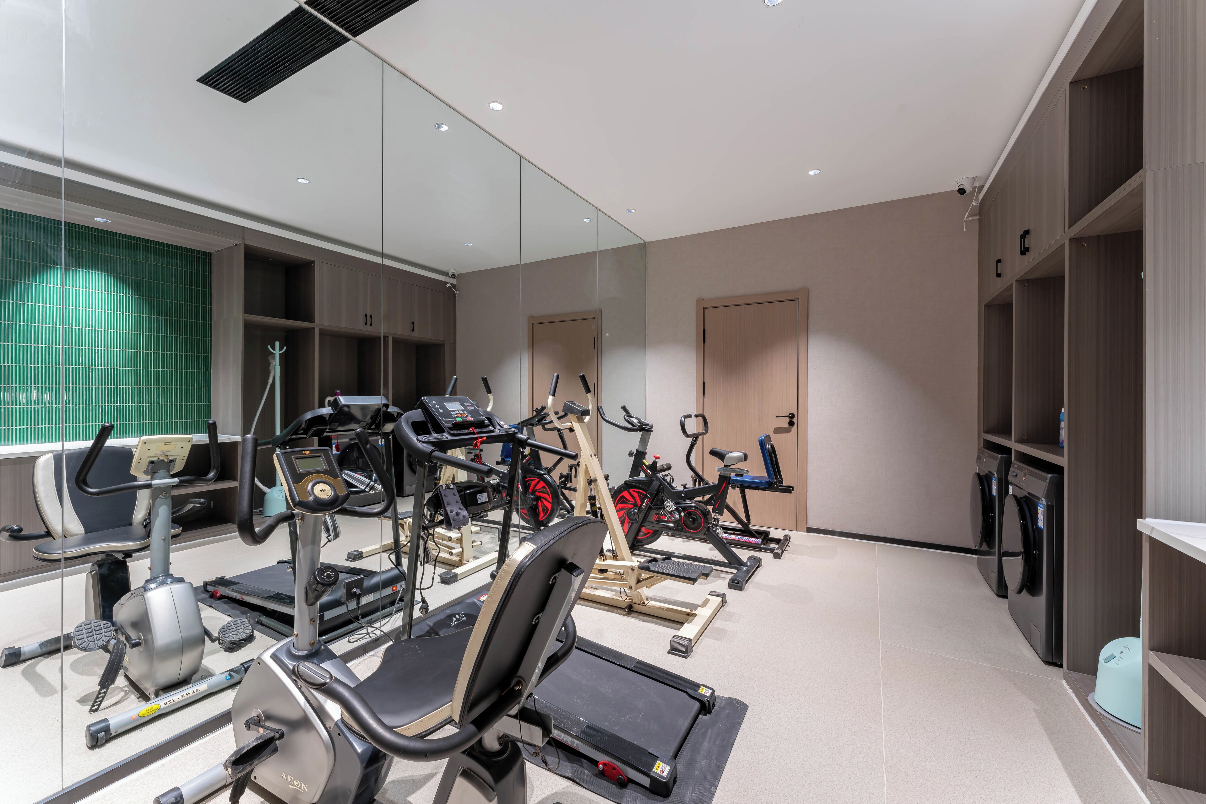 Une salle de sport moderne équipée de divers appareils de cardio, dotée d’un mur miroir et d’un sol carrelé et impeccable.
