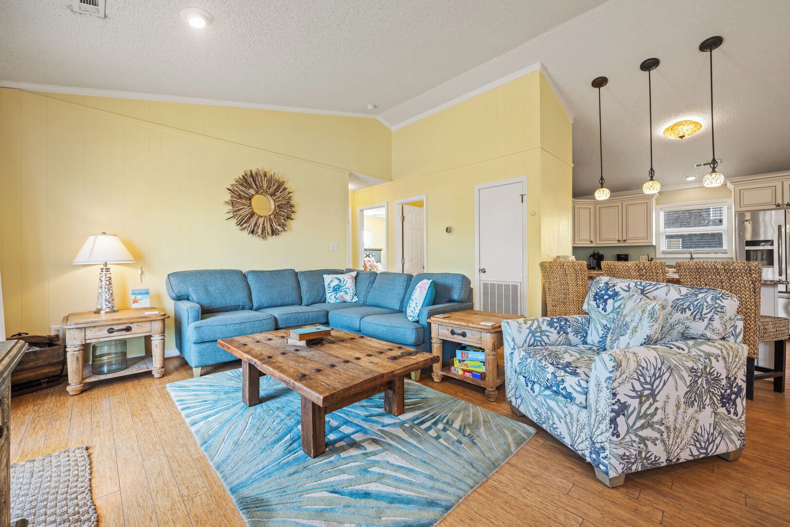 Ein Wohnzimmer mit einem blauen Modulsofa, einem Holztisch und einem gemusterten Sessel. Es gibt eine Küchenzeile mit weißen Schränken und Edelstahlgeräten.