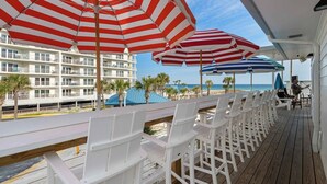 A deck with white plastic chairs and red and white striped umbrellas, overlooking a beach and a hotel building. - The Adventurer’s Micro Studio at Beachview (Fort Walton Beach)