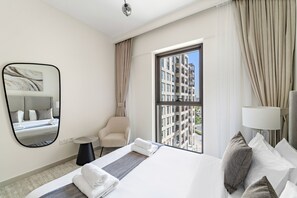 A hotel room with a large bed, a chair, a small table, a mirror, and a window with a view of buildings. - Bayshore at Creek Beach 3 - 501 1BD (Dubai)