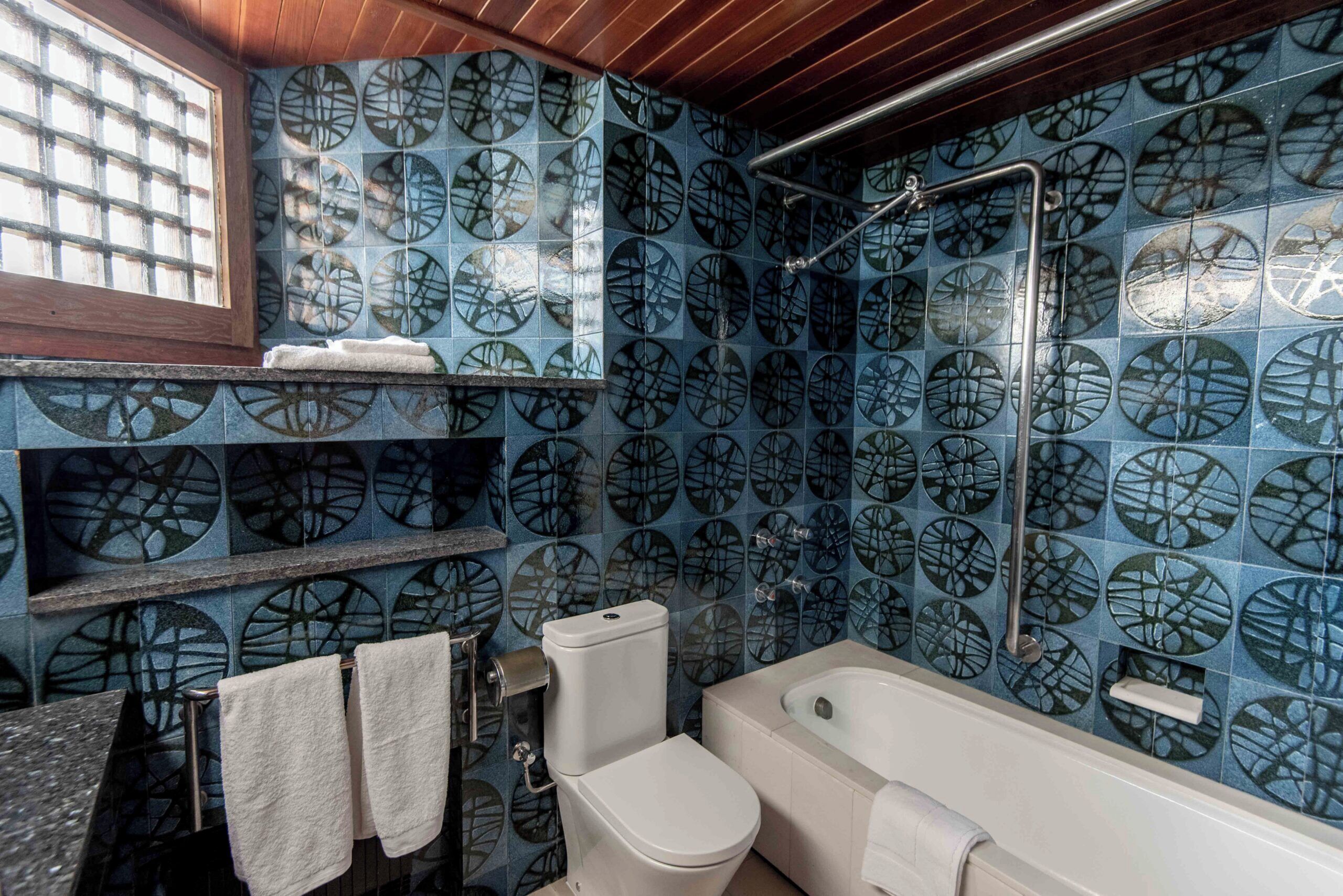 Une salle de bain avec un mur recouvert de carreaux bleus à motifs, des toilettes blanches, une baignoire et une fenêtre située au-dessus des toilettes.