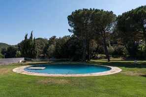 Une piscine ronde entourée d’une terrasse en pierre, elle-même bordée d’une pelouse verdoyante et d’arbres.