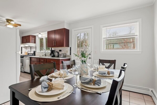 A dining area with a table set for four, kitchen cabinets, a window with blinds, and a ceiling fan.