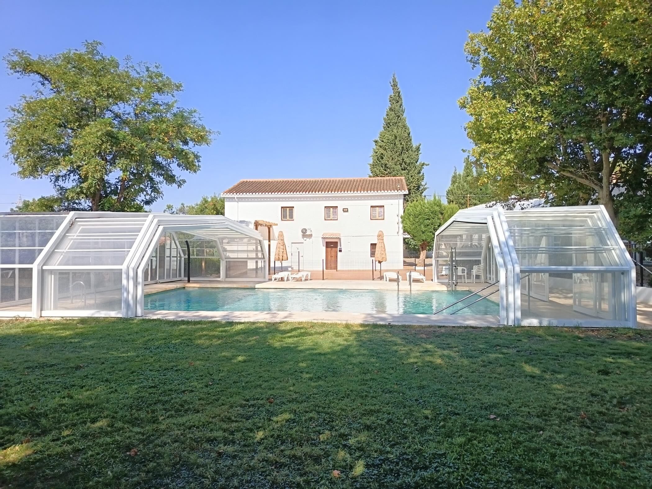 A swimming pool with glass-enclosed structures, a white building with a tiled roof, and surrounding trees.
