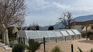 Estructura de invernadero con valla metálica, rodeada por una zona pavimentada con guijarros blancos y pequeñas palmeras.