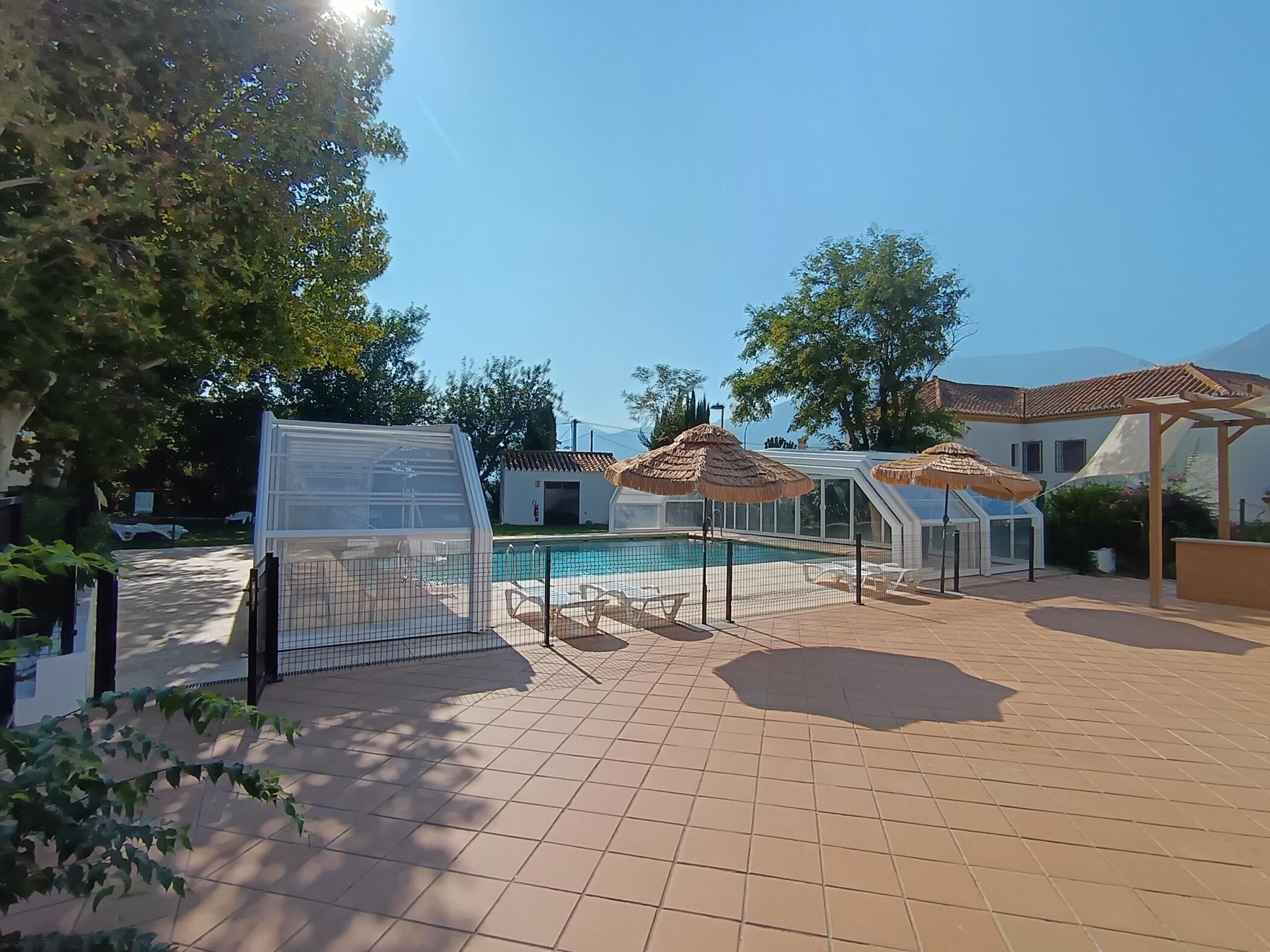 A swimming pool with a covered area, a fenced garden, and a building with a tiled roof.
