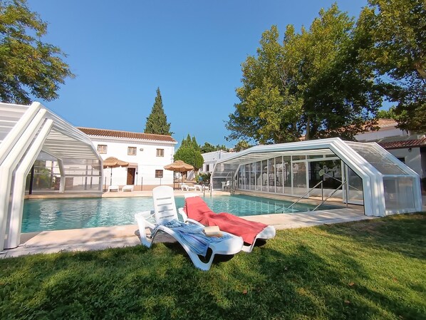 Une piscine dotée de chaises longues et d’un espace abrité, entourée de pelouse et d’arbres.