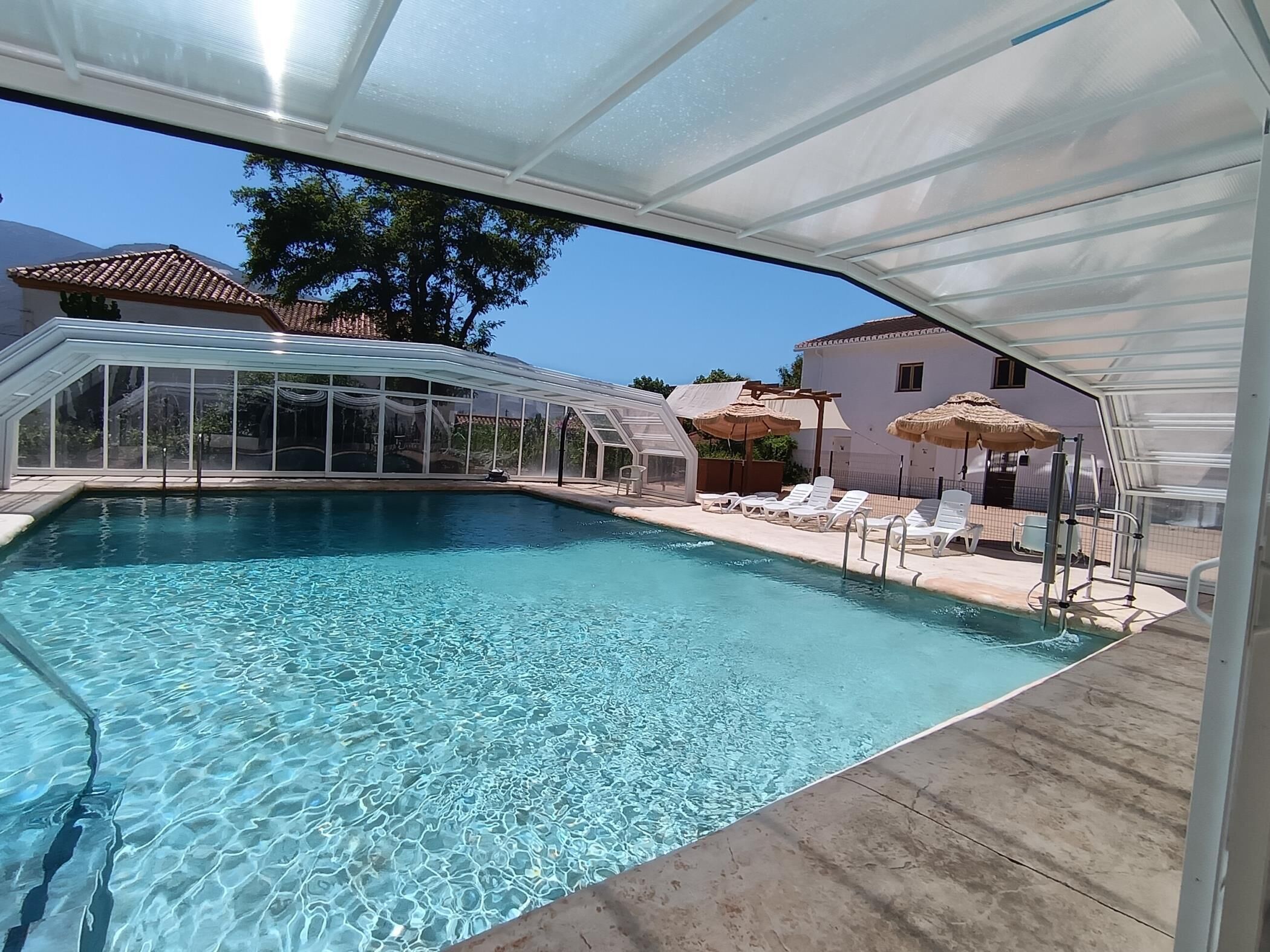 A clear swimming pool with lounge chairs and umbrellas, surrounded by a covered area with a glass roof.