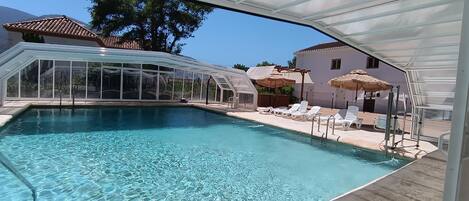 A clear swimming pool with lounge chairs and umbrellas, surrounded by a covered area with a glass roof.