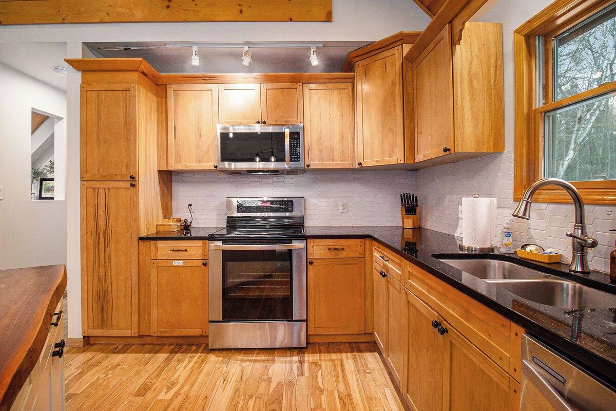 Une cuisine avec des meubles de rangement en bois, des appareils électroménagers en acier inoxydable, un plan de travail noir et une fenêtre.