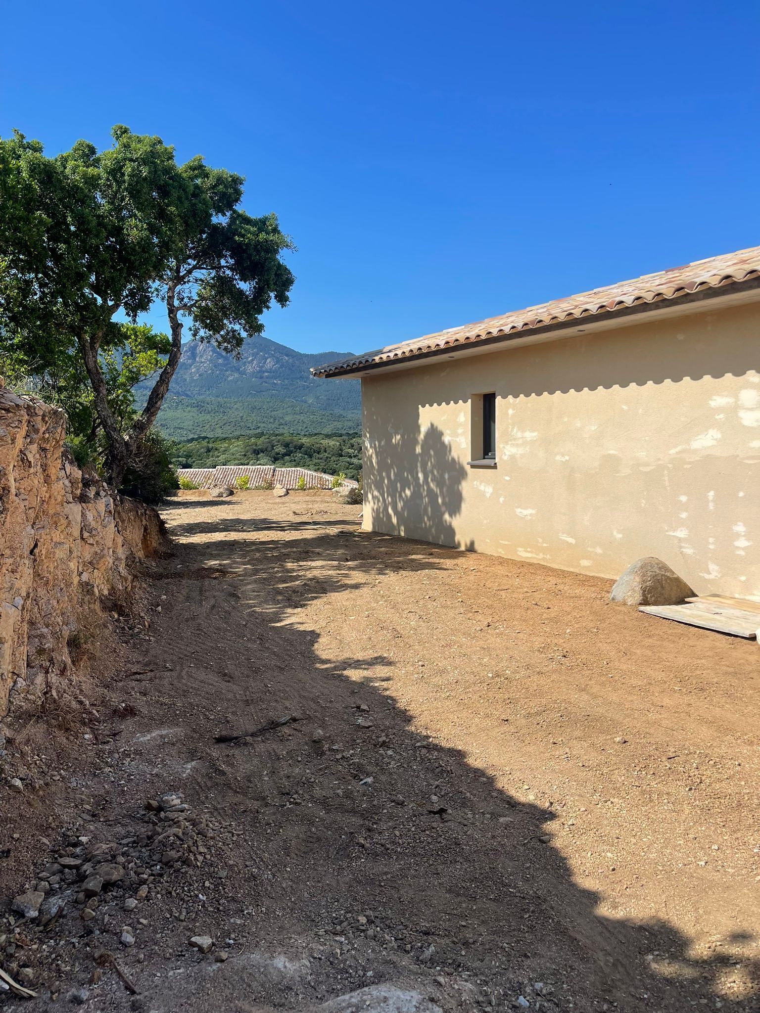 A rustic building with a dirt path leading to it, surrounded by trees and mountains in the background.
