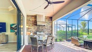 Une cuisine moderne avec un mur de crédence en pierre, un coin repas avec des meubles en osier et une terrasse fermée par des baies vitrées donnant sur un espace verdoyant.