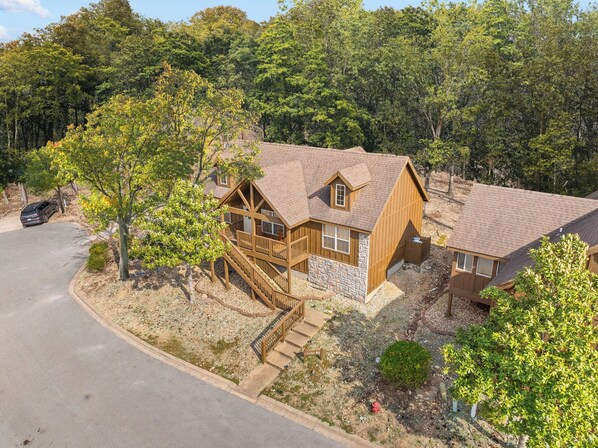 A two-story wooden cabin with a stone foundation, a balcony, and a driveway leading to a parked car.