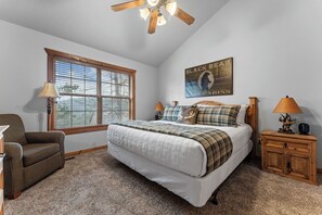 A bedroom with a large bed, two bedside tables, a chair, a window with blinds, and a ceiling fan.