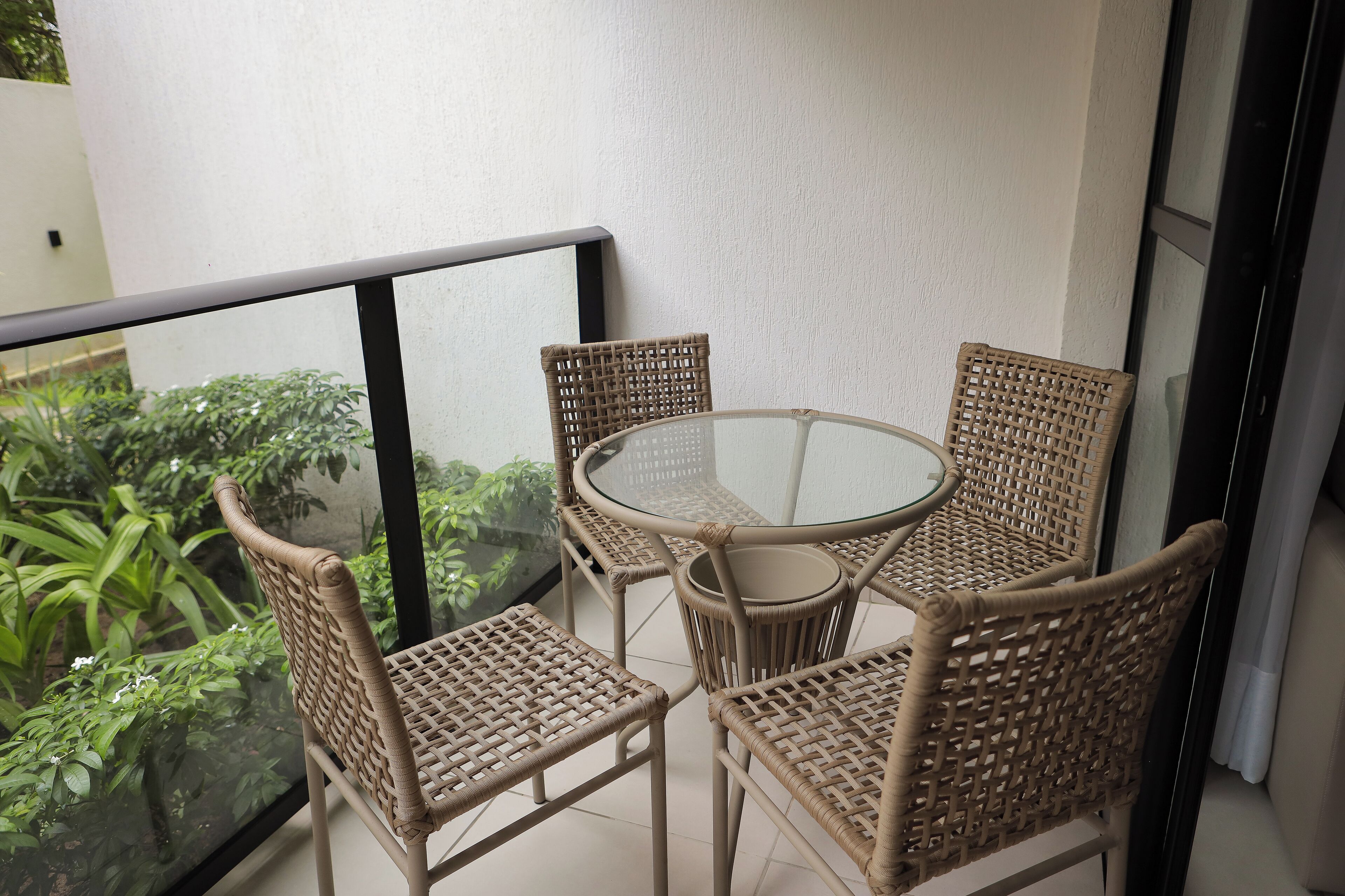 A balcony with wicker chairs and a glass table, overlooking a garden with green plants and white flowers.