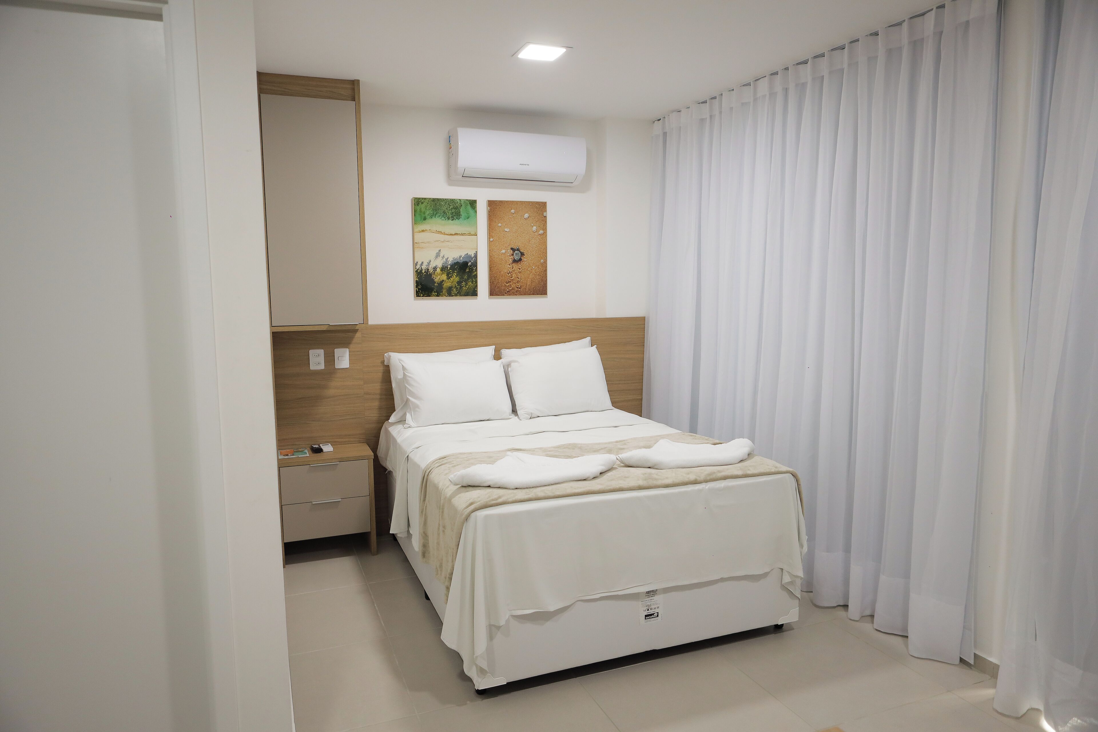Habitación de hotel con cama, mesita de noche, aire acondicionado de pared y dos obras de arte.