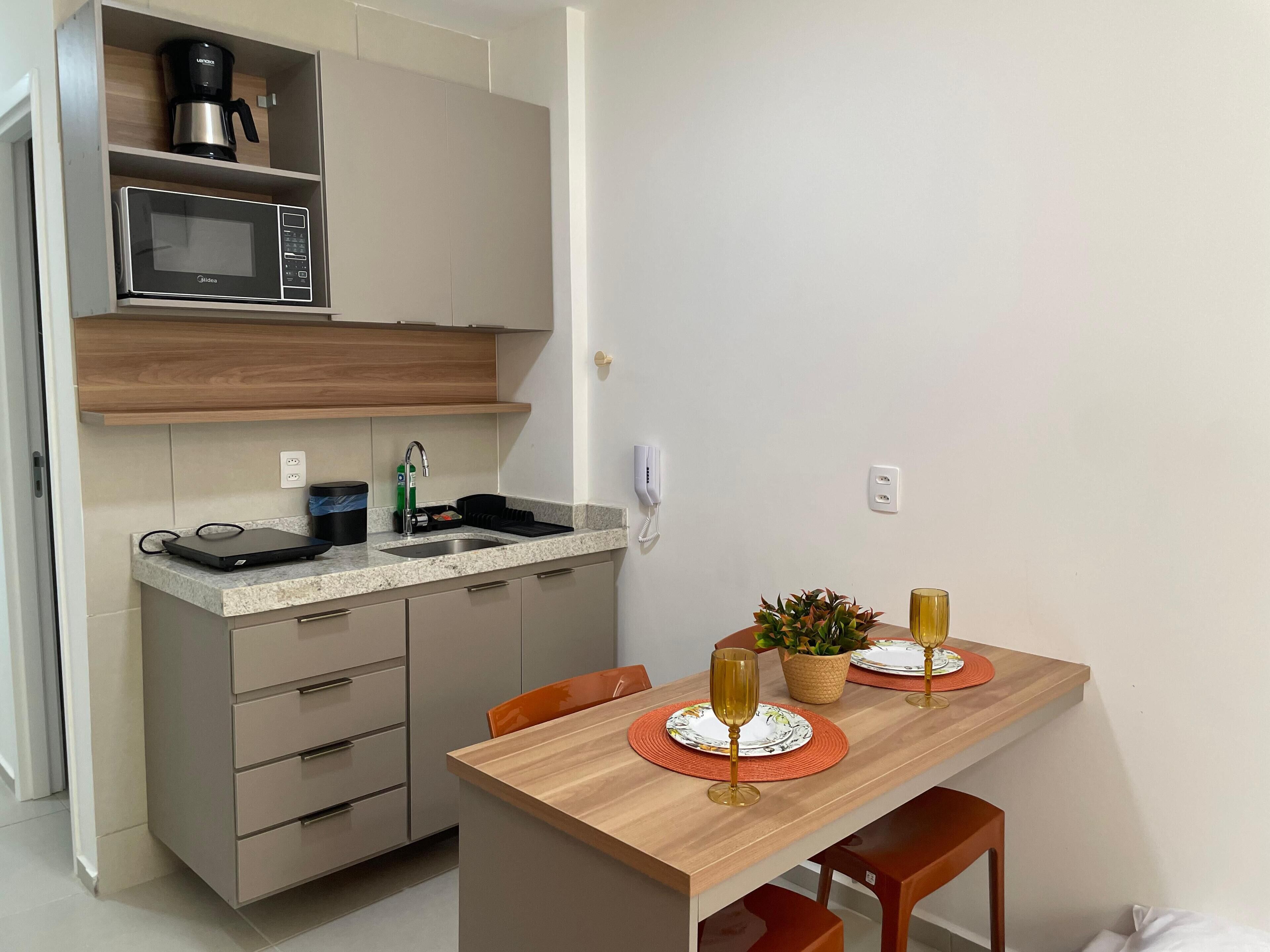 Una cocina compacta con microondas, cafetera y pileta. Hay una mesa de comedor con dos copas de vino y una planta en maceta.