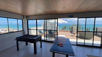 A spacious room with a foosball table and ping pong table, large windows offering ocean views, and a balcony with outdoor seating.