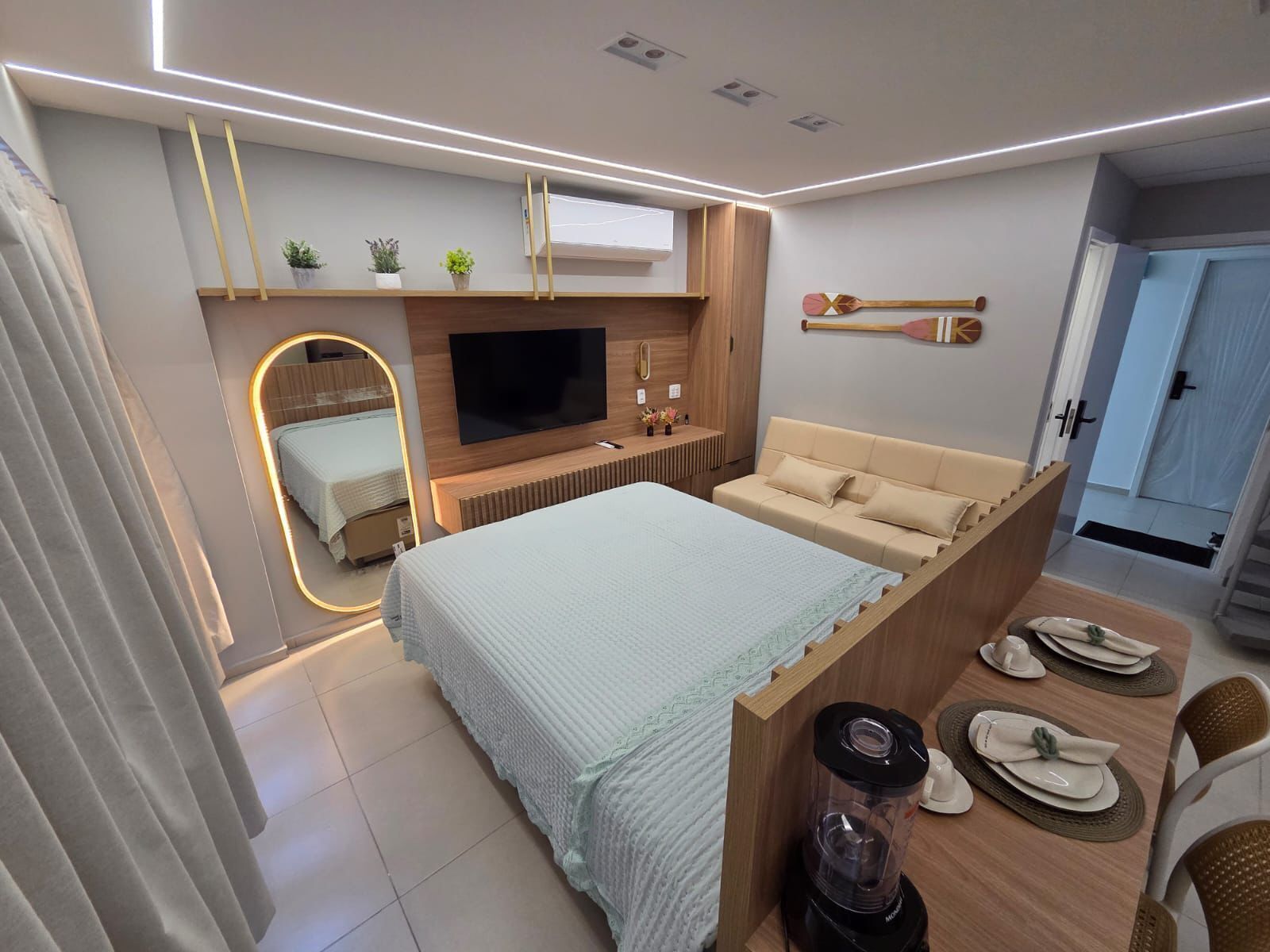 Ein modernes Schlafzimmer mit Bett, Fernseher, einer kleinen Sitzecke und einem Esstisch mit Tellern und einem Mixer.
