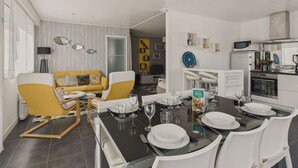 Uma sala de estar moderna com um sofá amarelo, uma mesa de jantar posta para seis pessoas e uma cozinha ao fundo.