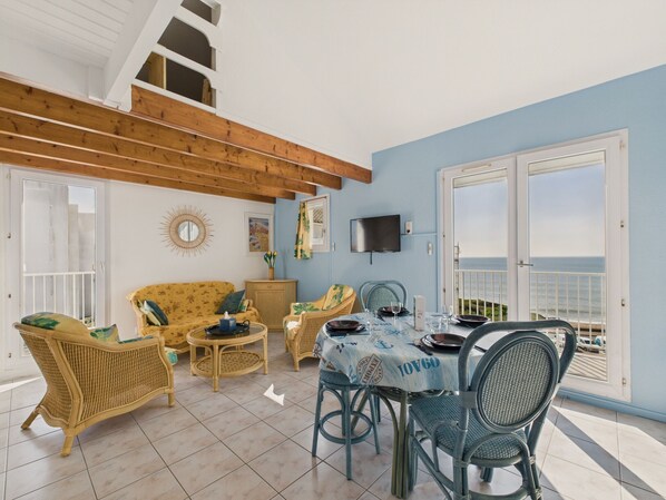 A dining area with a table set for four, surrounded by wicker chairs, a sofa, and a television mounted on the wall.