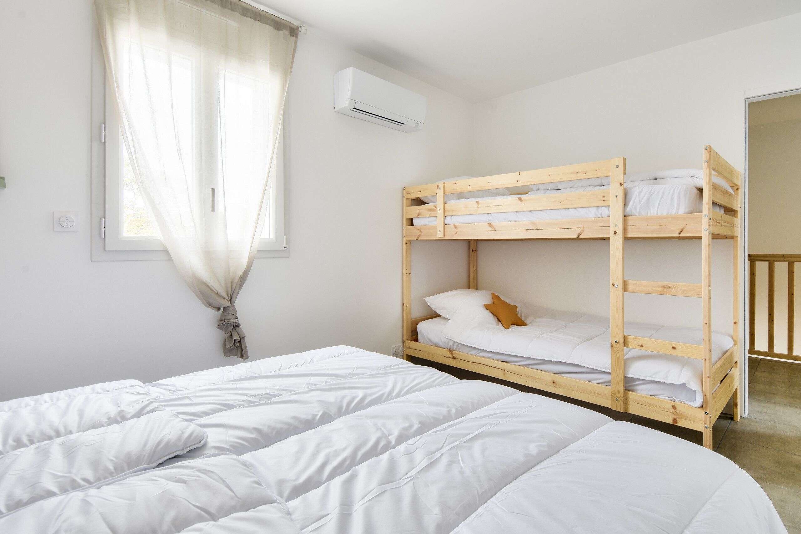 A bunk bed with white bedding, a window with sheer curtains, and an air conditioning unit on the wall.